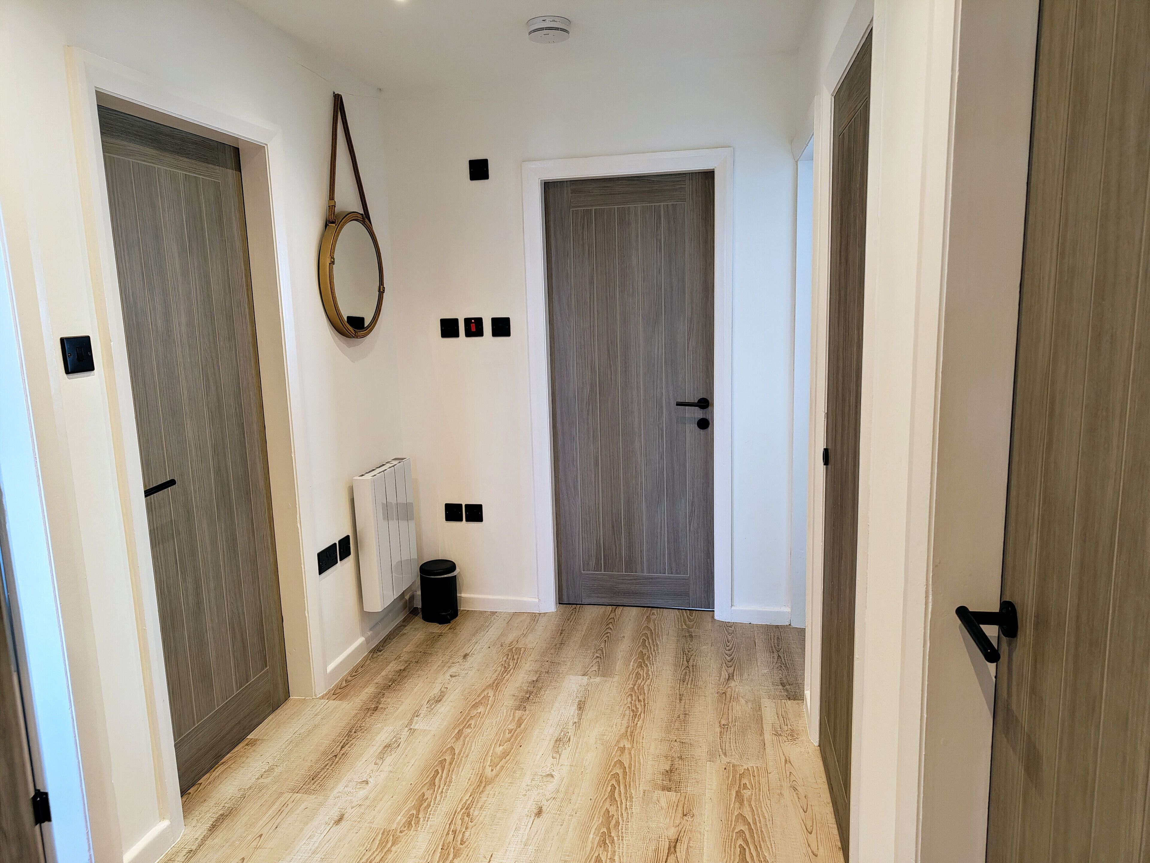 A hallway with wooden flooring, a white door with a black handle, and a mirror on the left wall.