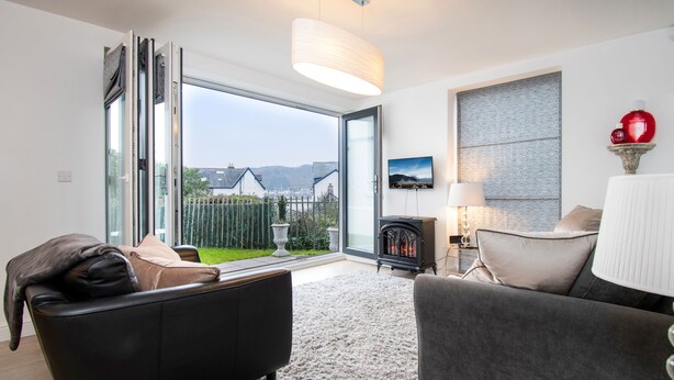 A modern living room with a large sliding glass door leading to a balcony, a fireplace, and a view of houses and mountains. - Eleven - One Bedroom Cottage, Sleeps 2 (Deganwy)