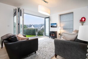 Un salon moderne doté d’une grande porte-fenêtre coulissante donnant sur un balcon, d’une cheminée et offrant une vue sur des maisons et des montagnes.