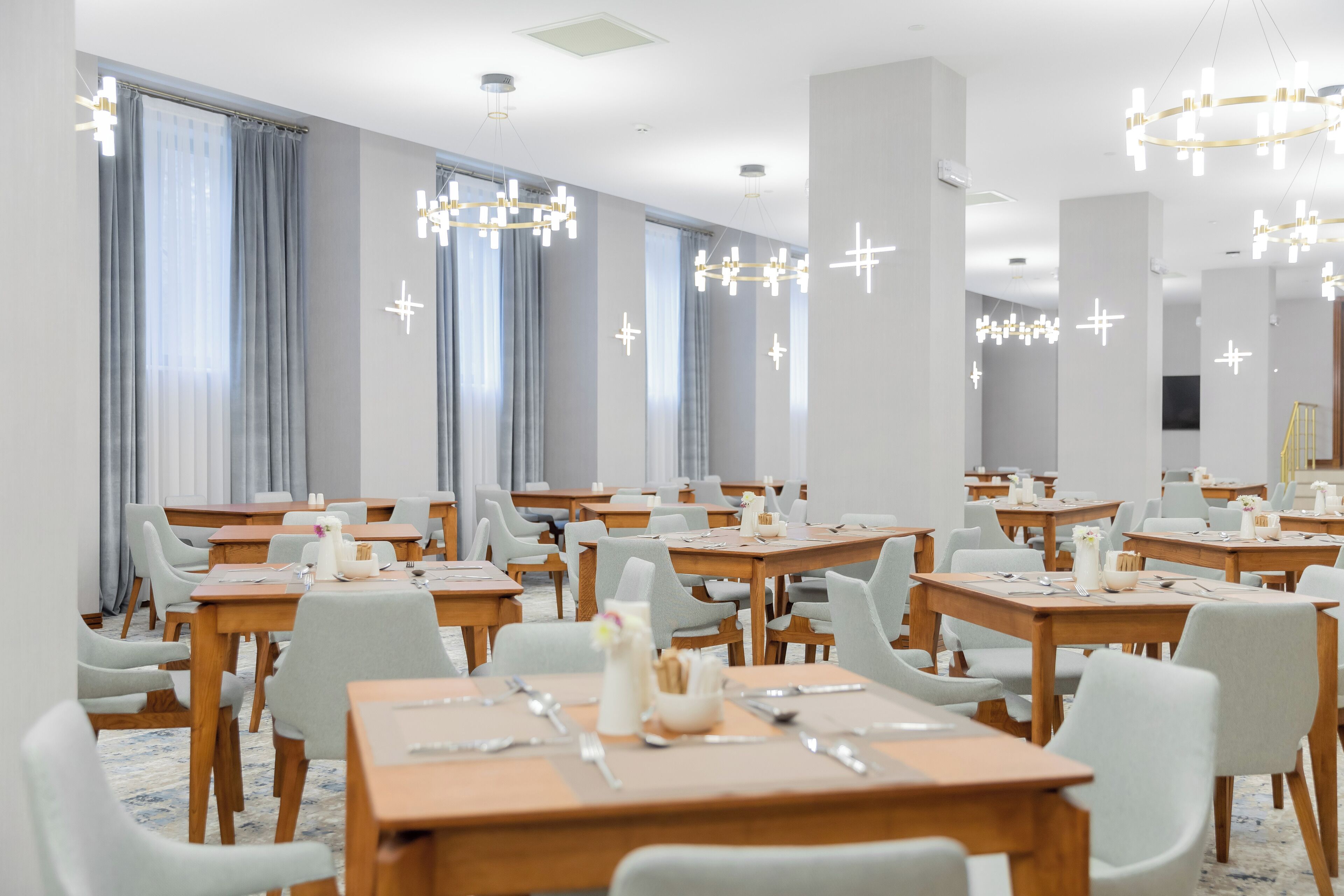 A hotel dining area with multiple tables and chairs, large windows with curtains, and modern chandeliers.