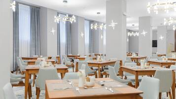 Un comedor de hotel con varias mesas y sillas, amplias ventanas con cortinas y candelabros modernos.
