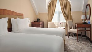 Ein Hotelzimmer mit zwei Betten, einem Schreibtisch, einem Stuhl, einem kleinen Tisch, einem Spiegel und einem Fenster mit Vorhängen.