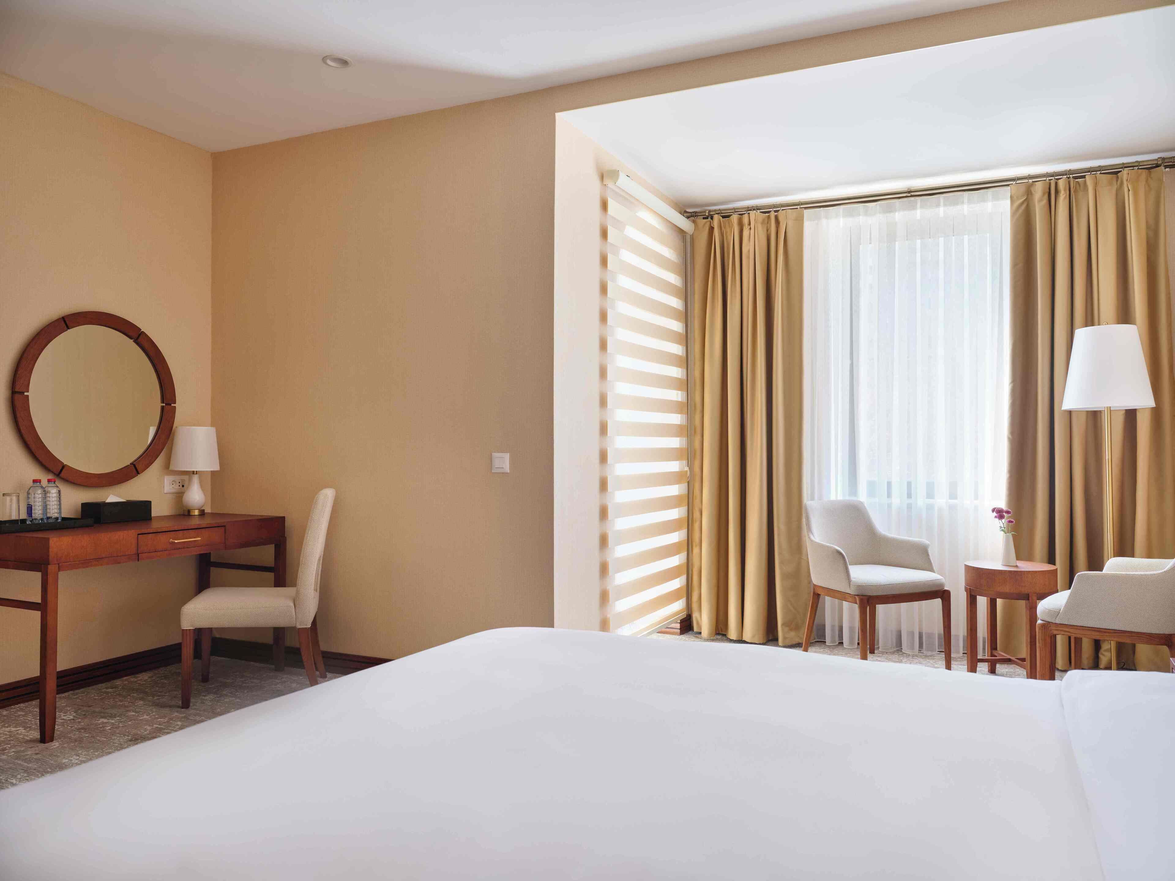 A hotel room with a bed, a desk with a chair, a round mirror, a lamp, and a window with curtains.