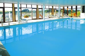 An indoor swimming pool with clear glass walls, multiple lounge chairs, and a view of the surrounding landscape. - Wooden Chalet in Abbeville near Somme Bay (Grand-Laviers)