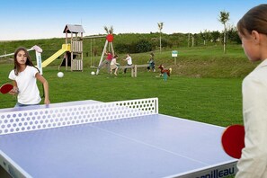 Una mesa de ping pong con una red blanca y, al fondo, un área de juegos con un tobogán y columpios.
