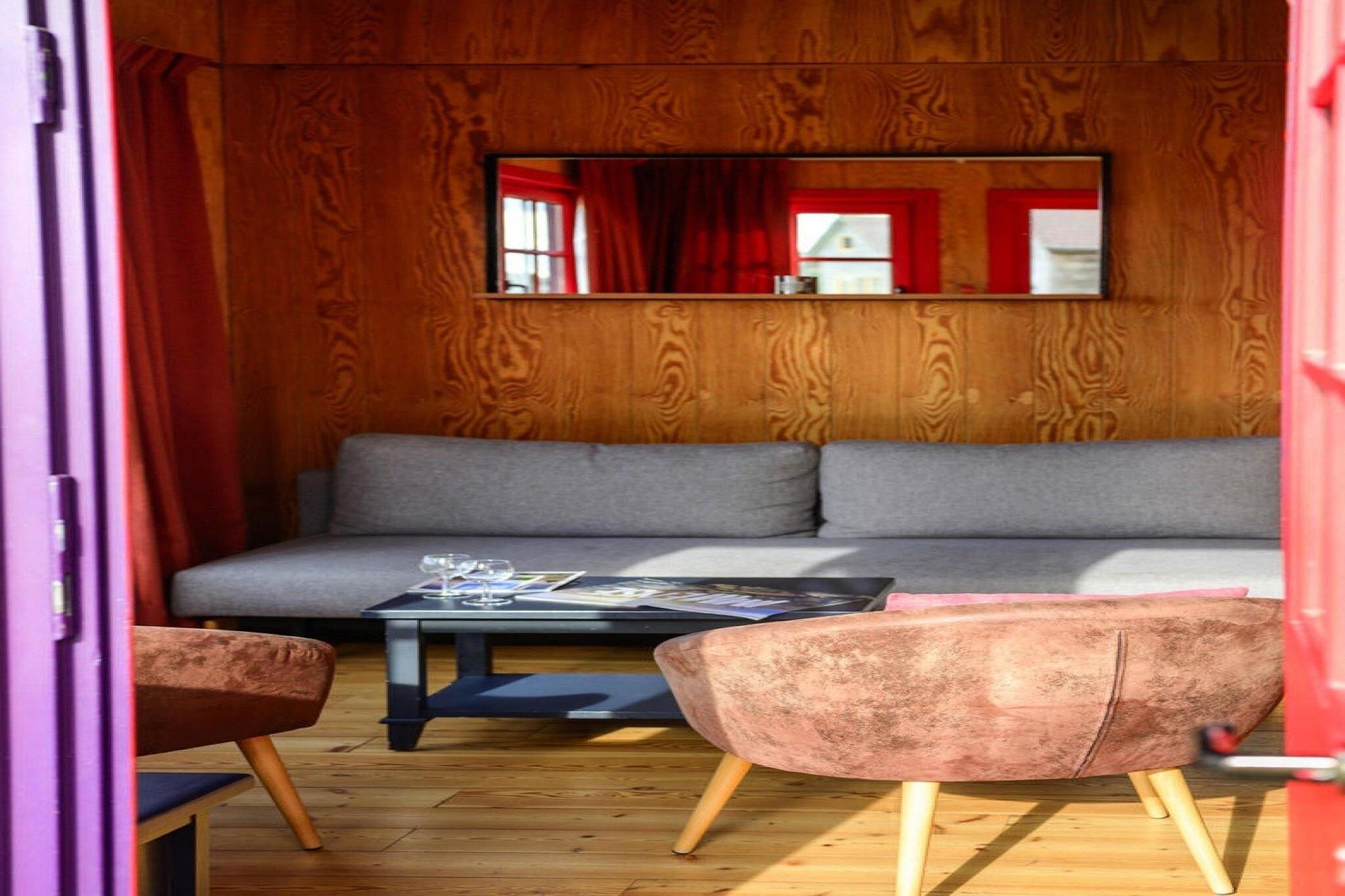Een gezellige kamer met een houten wand, een grijze bank, een klein tafeltje met glazen en twee stoelen.