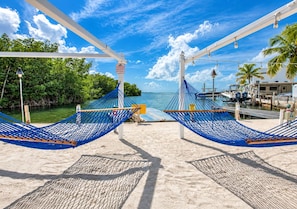 A beach view with two blue hammocks under a white canopy, a clear sky, and a marina with boats.