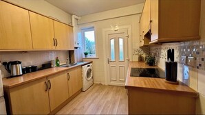 A compact kitchen with wooden cabinets, a washing machine, and a window with a view of greenery. - Holly street house with Separate Entrance, on the Edge of Glasgow
 (Clydebank)