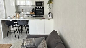 Ein modernes Wohnzimmer mit einem dunkelgrauen Sofa, einem Holztisch und einer Küchenzeile mit weißen Schränken, einem schwarzen Backofen und einer kleinen Pflanze auf der Arbeitsfläche.