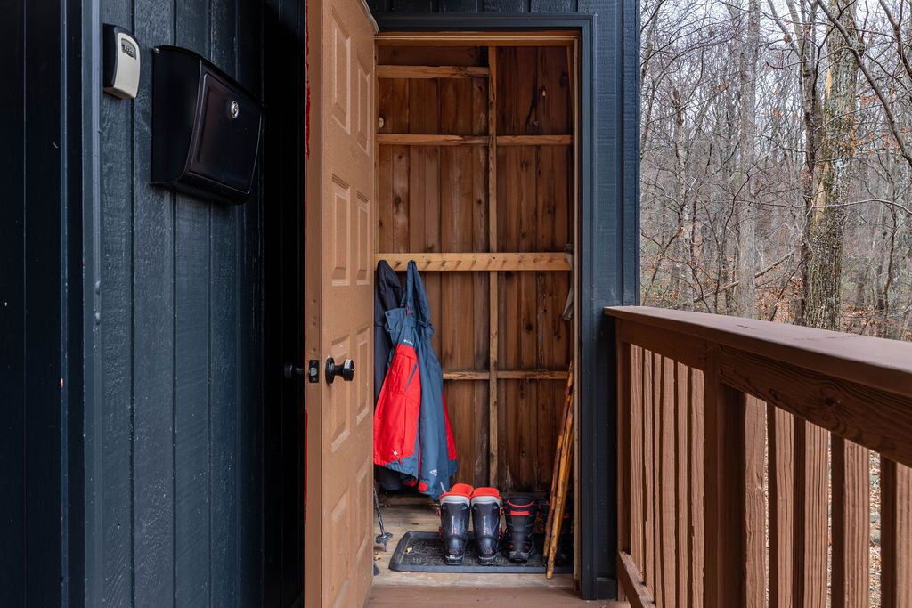 An open wooden door leading to a storage area with a bench, a coat rack with jackets, and a pair of skis and boots.