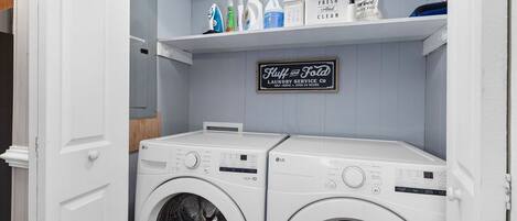 Ein Waschraum mit zwei LG Frontlader-Waschmaschinen, einem Regal mit Waschmitteln und einem Schild für 'Huff and Fold Laundry Service Co'.