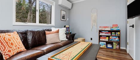 A living room with a brown leather sofa, a coffee table with a board game, a bookshelf with board games, and a window with a view of trees.