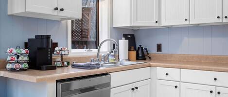 A modern kitchen with white cabinets, a stainless steel dishwasher, a coffee maker, and a window with a view.