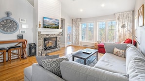 A cozy living room with a fireplace, a large clock, and a view of the outdoors through large windows. - Hawks Peak 406- Mtn Condo, Top Floor, Mtn View, Modern, Near Attractions (Seven Devils)