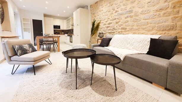 Ein modernes Wohnzimmer mit einem grauen Sofa, einem beigen Sessel und zwei runden schwarzen Tischen. Im Hintergrund ist eine Steinwand sowie eine Küche mit weißen Schränken zu sehen.