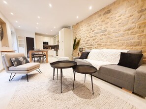 A modern living room with a grey sofa, a beige armchair, and two round black tables. A stone wall and a kitchen with white cabinets are visible in the background. - Voltaire's home in the heart of the city (Saint-Germain-en-Laye)
