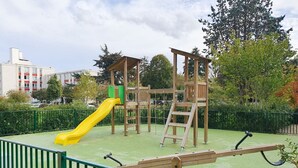 Ein Spielplatz mit einer gelben Rutsche und Klettergerüsten, umgeben von einem grünen Zaun und Bäumen.