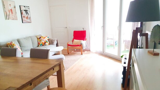 Ein großzügiger Wohnbereich mit einem Holzesstisch, einem roten Sessel und einer Couch. Ein großes Fenster lässt Tageslicht herein und bietet einen Blick ins Freie.