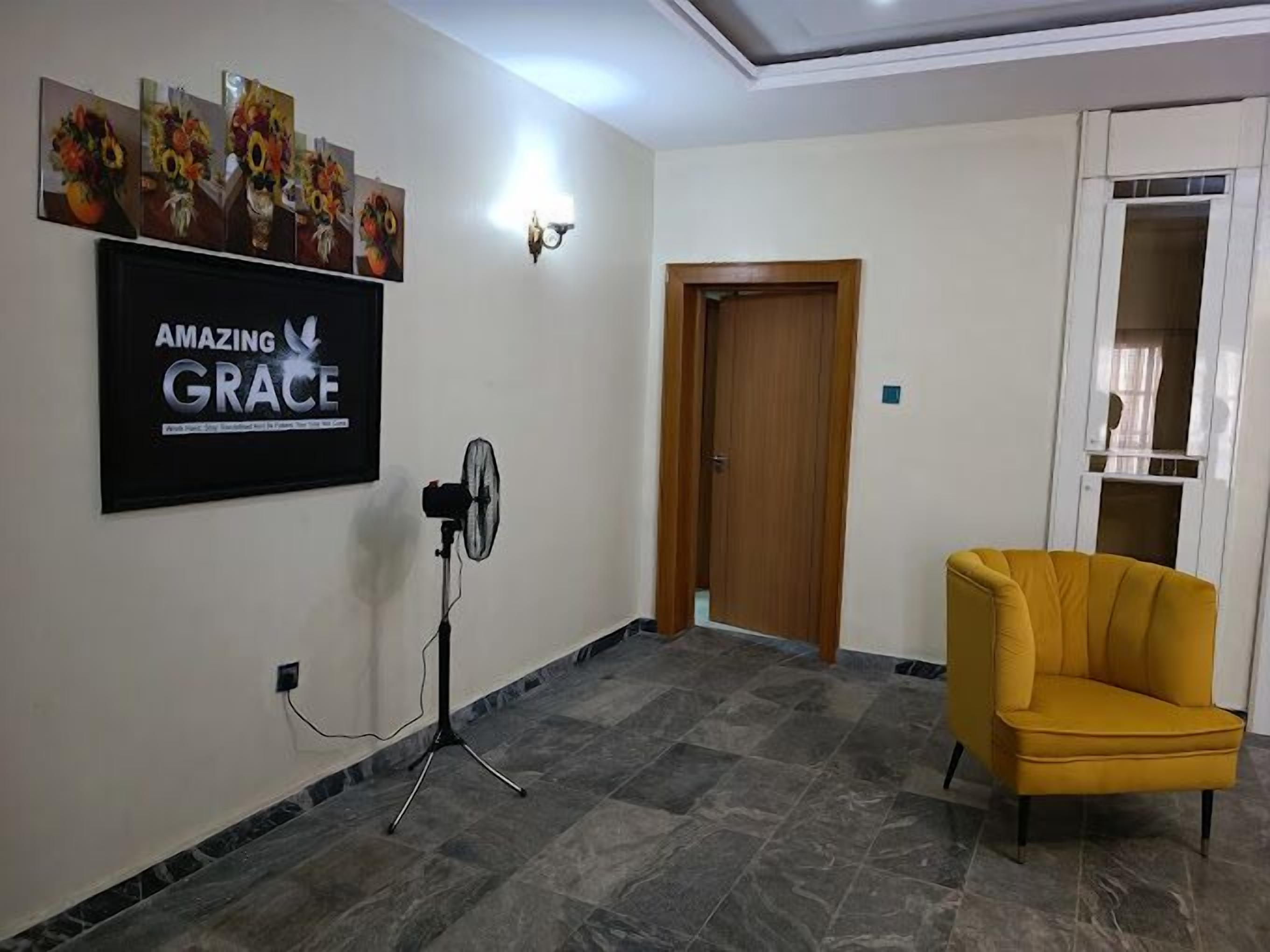 A room with a wooden door, a yellow armchair, a wall-mounted fan, and a framed artwork titled "AMAZING GRACE".