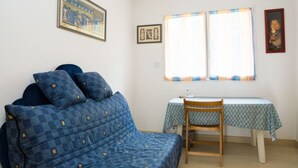 Una habitación pequeña y acogedora con un sofá azul estampado, una silla de madera, una mesita con mantel estampado y dos ventanas con cortinas.