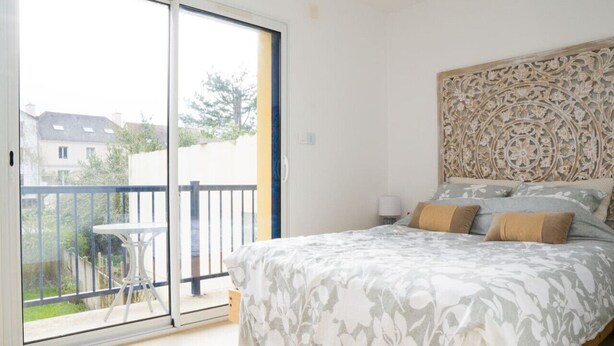 Un dormitorio con una cama grande, un cabecero decorativo, una mesita y un balcón con vistas a casas y vegetación.