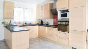 Una cocina moderna con armarios de madera clara, una isla central, electrodomésticos de acero inoxidable y un ventanal con cortinas.