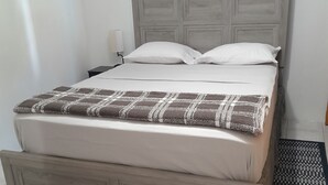 Un lit avec une tête de lit en bois, une literie blanche et un plaid à motifs. Un petit tapis à motifs géométriques est posé sur le sol.