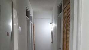 Un couloir étroit aux murs blancs et au sol carrelé, qui mène à une porte surmontée d’un volet en bois.