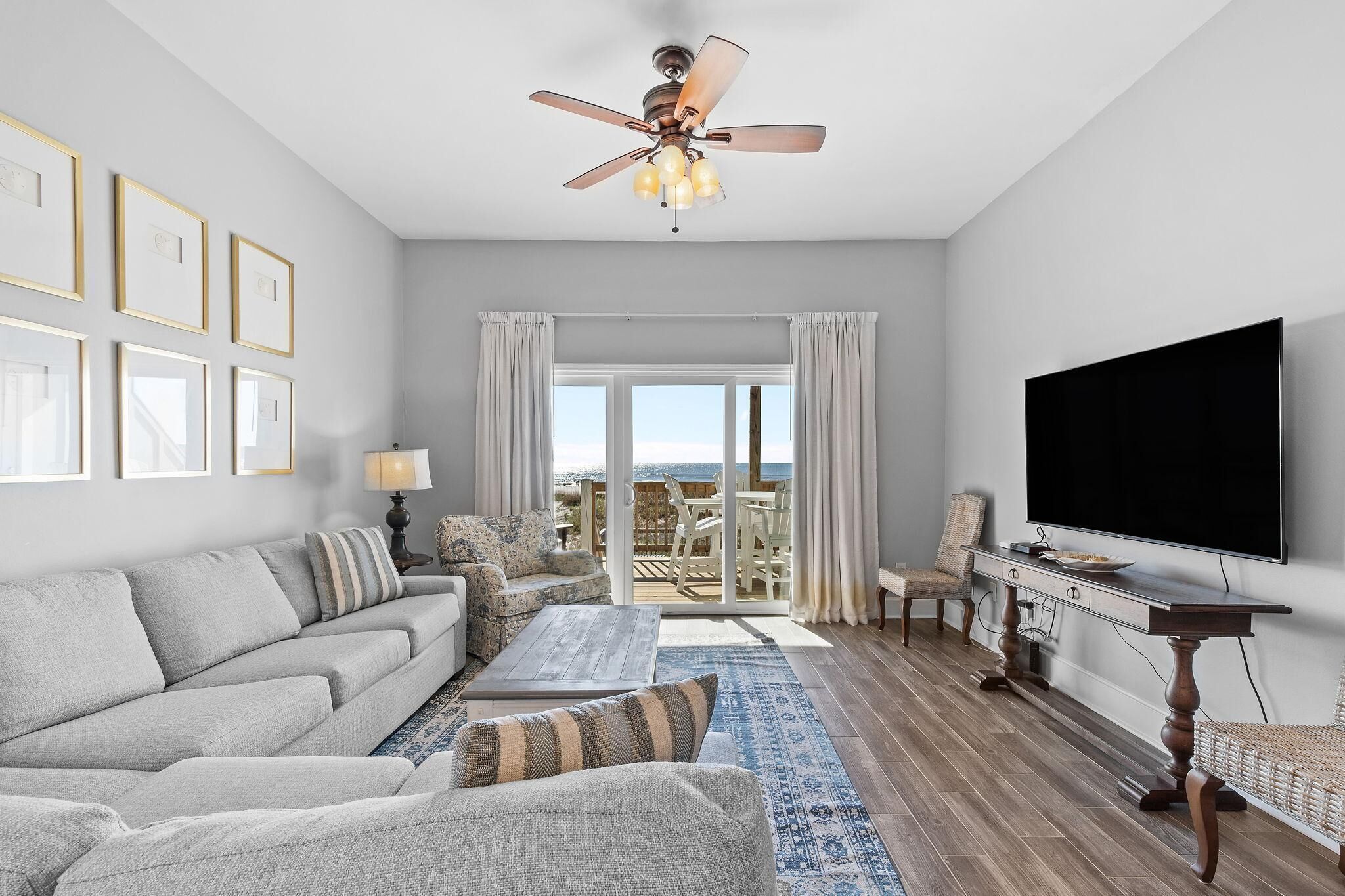 A spacious living room with a grey sofa, a coffee table, a TV, and a ceiling fan. There is a large window with curtains and a view of the outdoors.