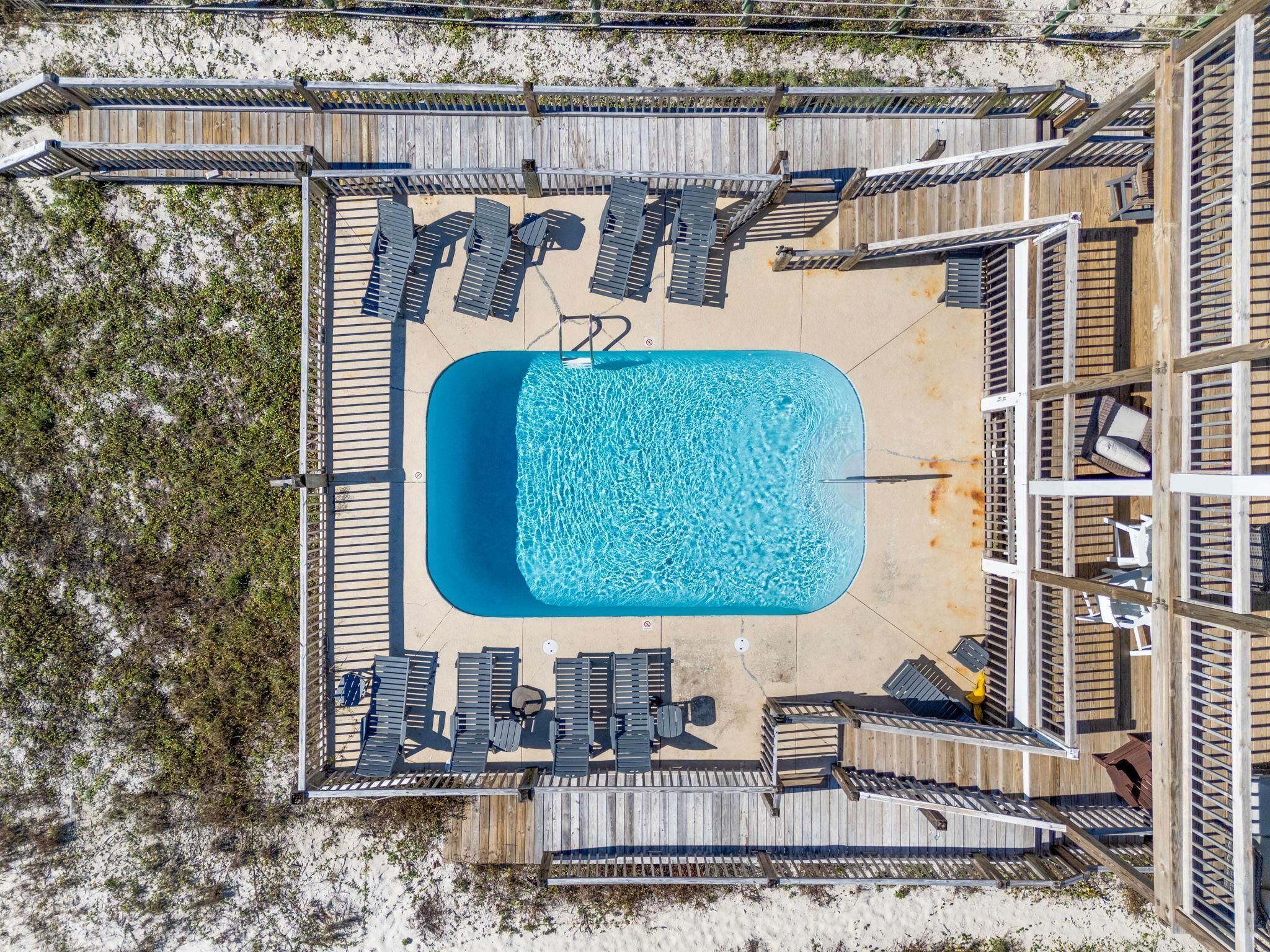 An aerial view of a beachside pool area with a rectangular pool, wooden deck, and lounge chairs.
