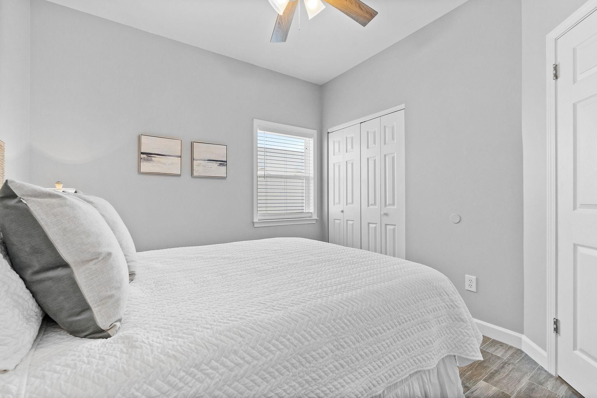 A bedroom with a bed, two wall-mounted picture frames, a window with blinds, a ceiling fan, and a white door.