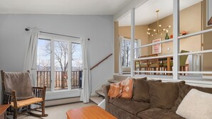 A living room with a sofa, a wooden rocking chair, a dining area with a table and chairs, and a staircase. - Sugarbush Condo | Hike, Bike & Explore (Warren)