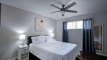 Une chambre à coucher comprenant un lit, une table de chevet, un ventilateur de plafond, une fenêtre avec des rideaux et une porte.