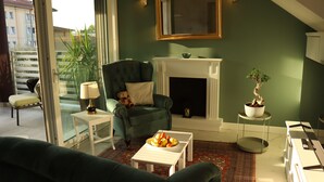 Un salon avec un fauteuil vert, une table basse blanche ornée de fruits, une cheminée et une vue sur un balcon agrémenté de plantes.