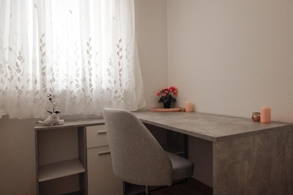 A minimalist desk with a grey chair, a lace window treatment, and a small vase with flowers.