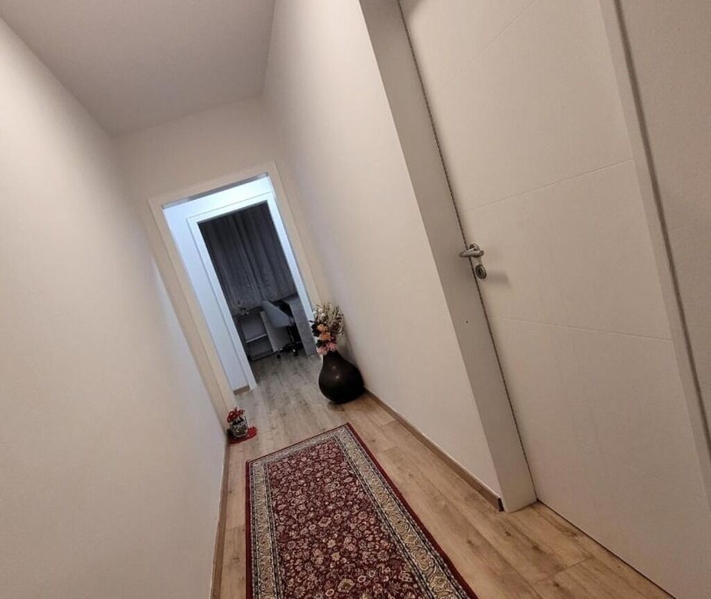 Un pasillo estrecho con piso de madera, un tapete estampado, una puerta con cerradura y un florero con flores en el suelo.