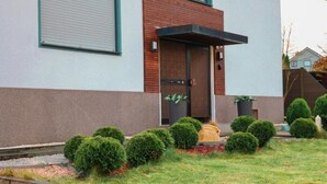Un edificio moderno con techo plano, un gran ventanal y una puerta con un pequeño alero. Se observan arbustos bien recortados y un pequeño jardín con un sendero.