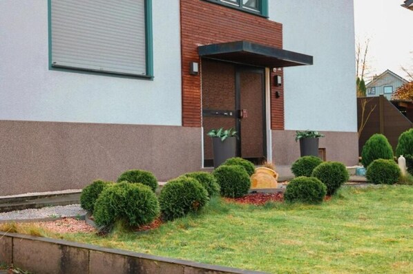 Ein modernes Gebäude mit flachem Dach, einem großen Fenster und einer Tür mit kleinem Vordach. Gepflegte Büsche und eine kleine Gartenanlage mit einem Weg sind zu sehen.