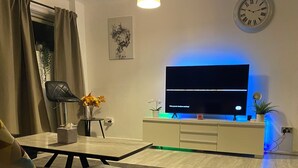 A modern living room with a flat-screen TV mounted on a wall, a small side table with a vase of flowers, a black chair, and a dining table with a vase of yellow flowers.