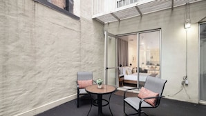 A small patio area with two chairs and a table, a sliding glass door leading to a bedroom, and a concrete wall. - Beautiful Studio with Patio in Midtown NYC! #202 (New York)