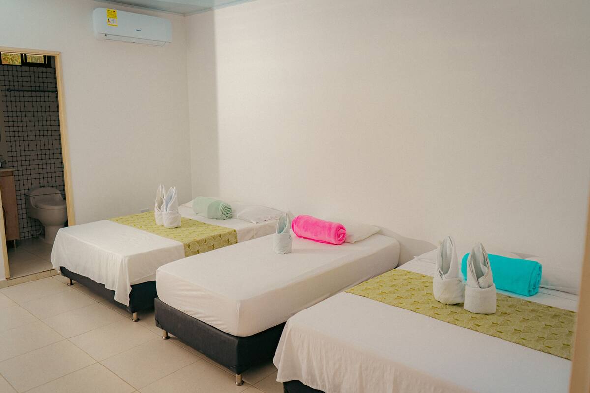A hotel room with three single beds, each with a white bedspread and a towel. There is a small window on the left wall, and a bathroom with a toilet and sink is visible through an open door.