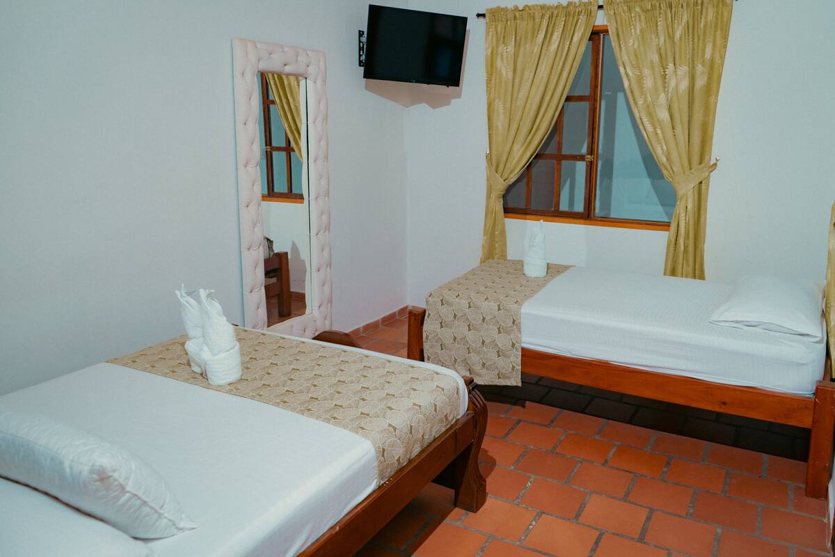 A room with two single beds, a TV mounted on the wall, and a window with curtains.
