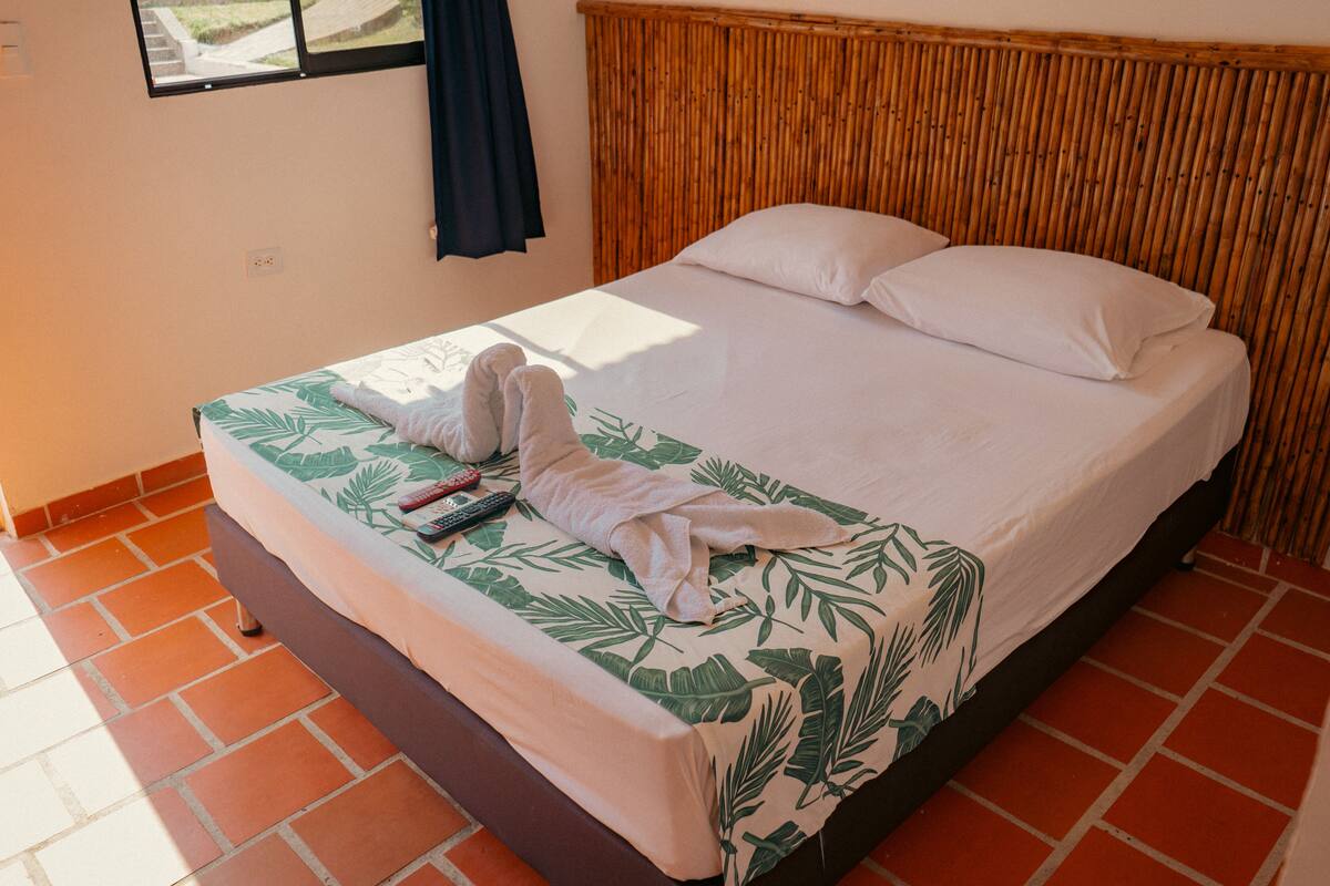 A neatly made bed with a tropical leaf pattern bedspread, a pair of white bathrobes, and two remote controls on the bed.