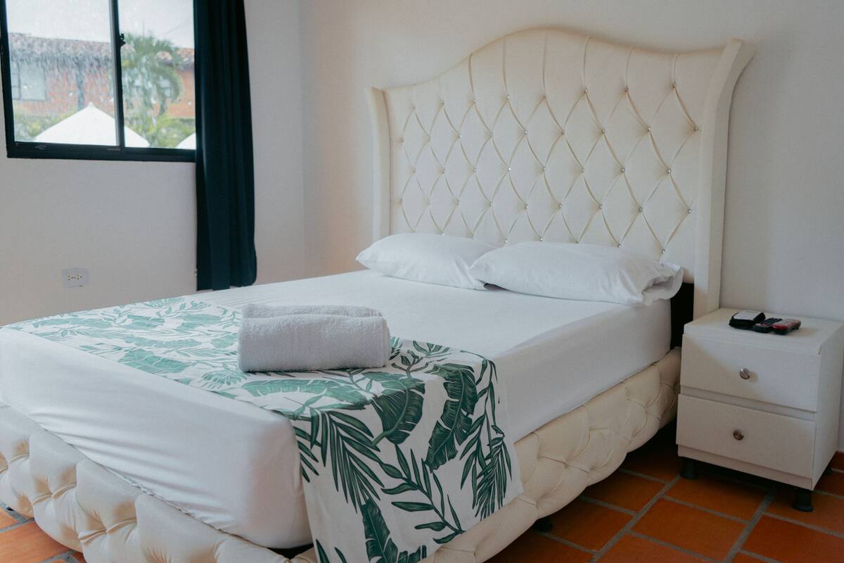 A neatly made bed with a white and green leaf-patterned bedspread, a white headboard, and a small bedside table with a drawer.