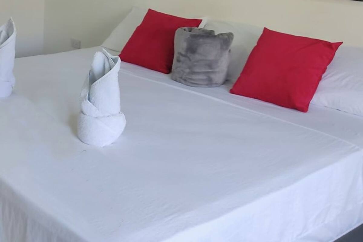 A neatly made bed with white linens, red pillows, and two rolled towels.
