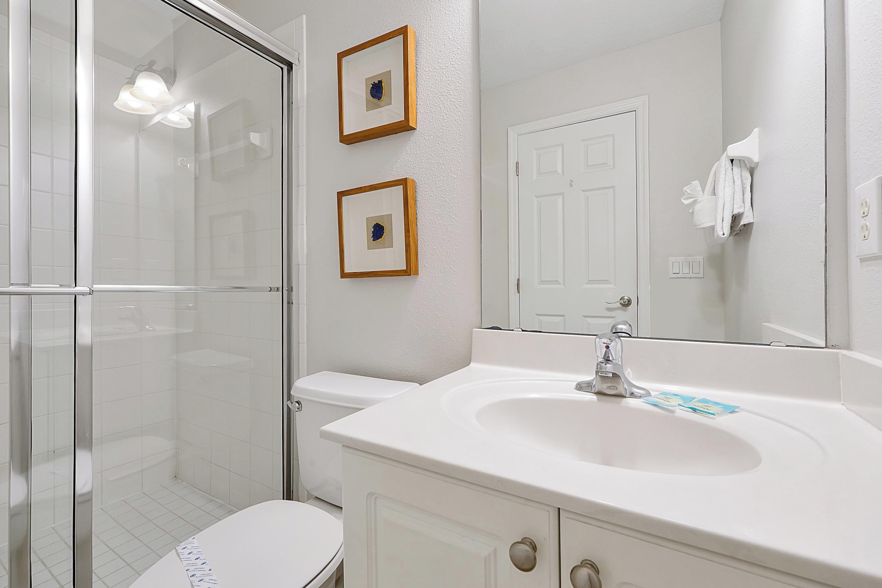 Une salle de bain avec une douche à parois de verre, des toilettes blanches, un lavabo avec un miroir et deux tableaux encadrés au mur.