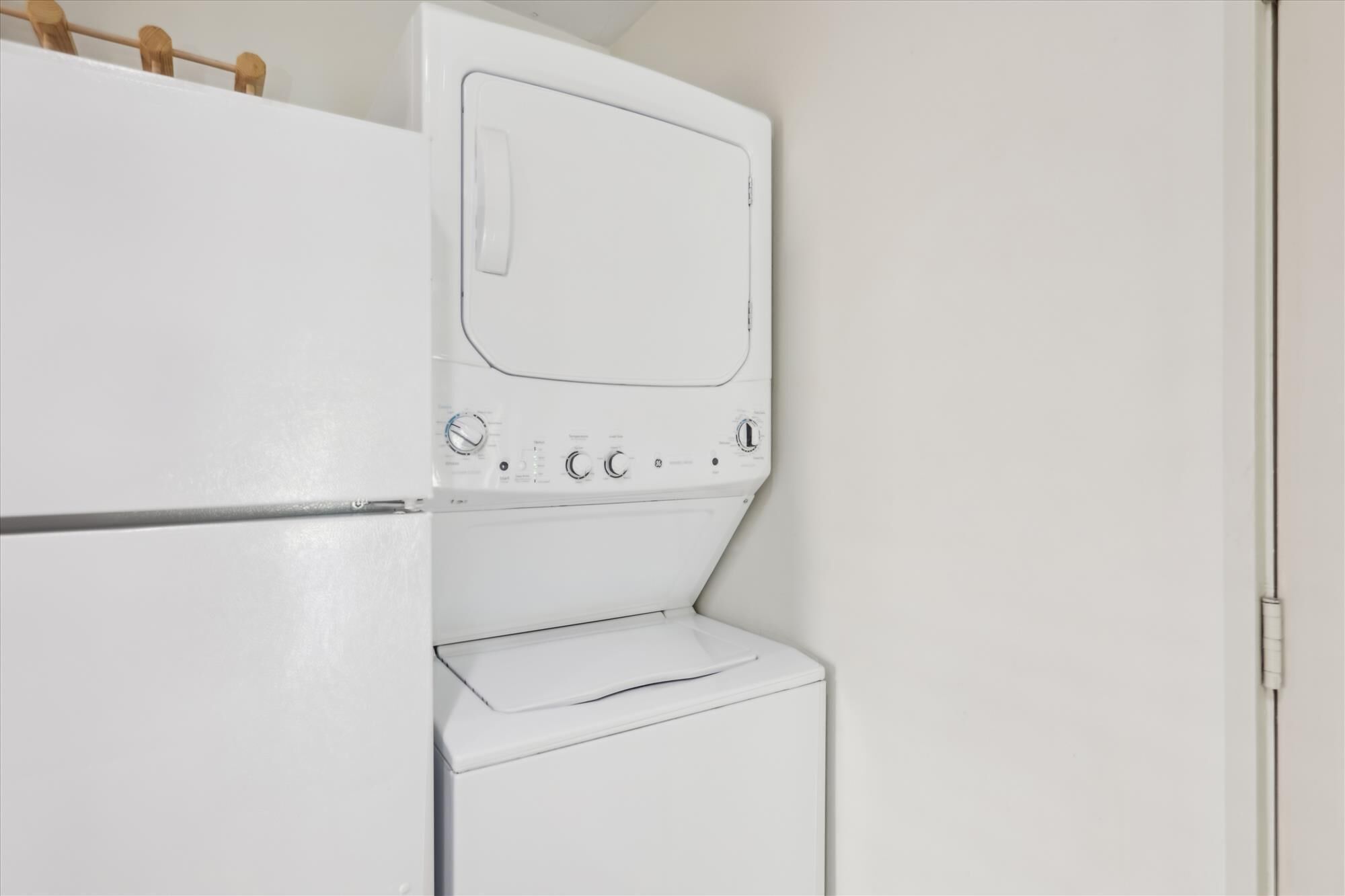 A compact, white laundry room with a built-in washer and dryer, a small countertop, and a wooden shelf above the dryer.