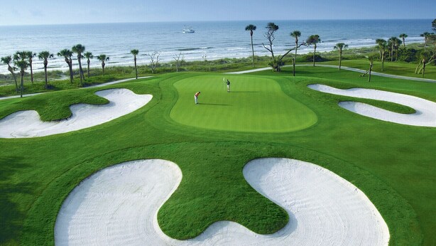 A golf course with sand bunkers, a person playing golf, and a view of the ocean.