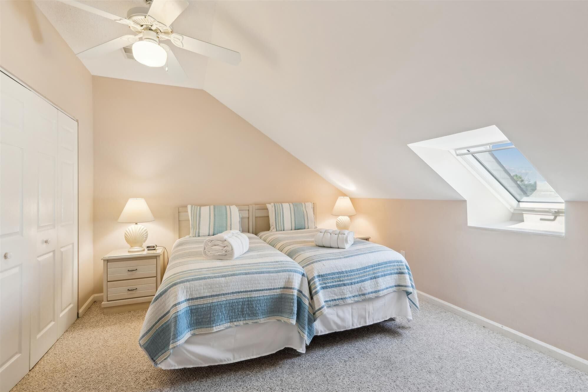 A bedroom with two beds, a ceiling fan, a skylight, and a nightstand with a lamp.