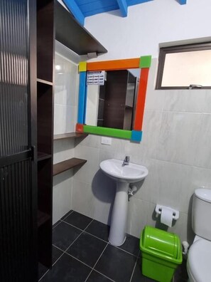 A bathroom with a white sink, a green trash bin, a toilet, and a mirror with a colorful frame. - Hotel Acuarela del Rio (Santa Fe de Antioquia)